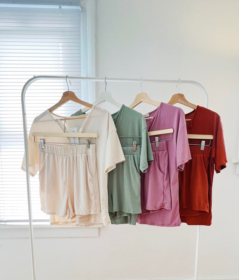 May include: Four sets of matching pajamas, each with a short-sleeved top and shorts. The pajamas are displayed on hangers and are in the colors beige, green, purple, and rust.