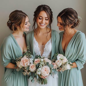 May include: Three women in sage green bridesmaid dresses with a white lace wedding dress in the center. The women are holding a bouquet of blush pink and white roses with greenery.