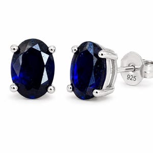 May include: A pair of oval-cut deep blue sapphire-colored stud earrings set in silver prongs. The earring backs are clearly marked with the number 925, indicating sterling silver construction. The stones feature a faceted surface and a dark blue hue.