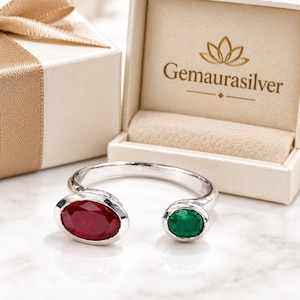 May include: A silver open-style ring featuring one large oval-cut red gemstone and one smaller round-cut green gemstone. The ring is positioned in front of an open cream-coloured jewellery box with the text Gemaurasilver printed on the inside lid.