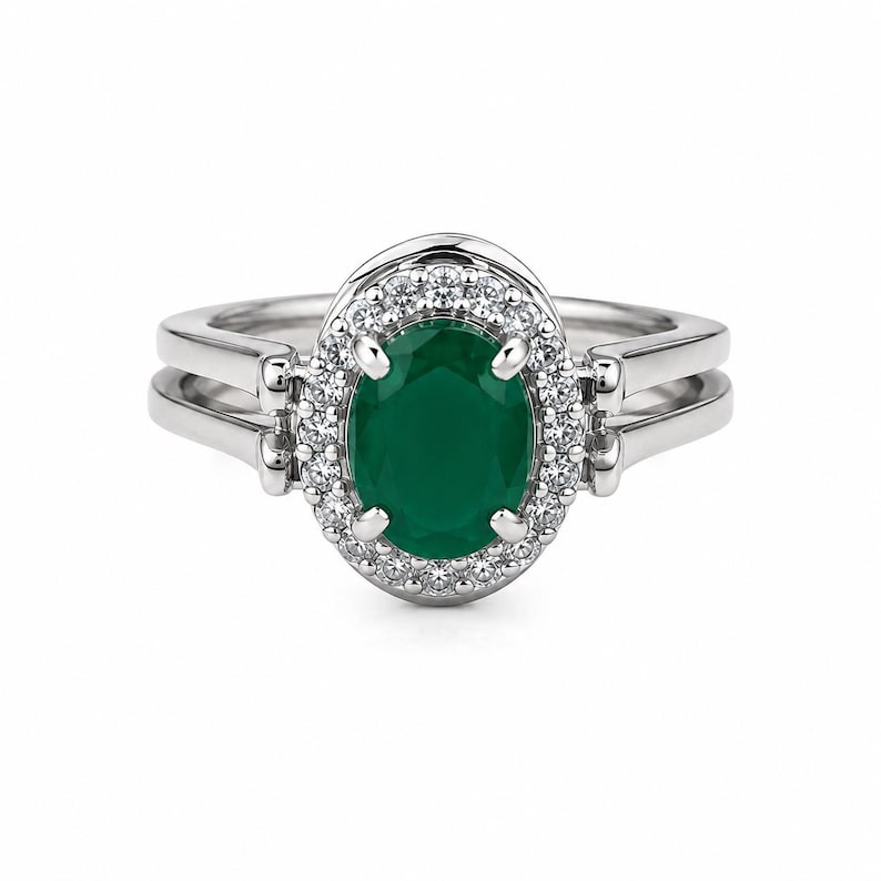May include: A silver-toned ring featuring a central oval-cut dark green gemstone surrounded by a halo of small clear stones. The band is split into two parallel sections on each side, connecting to the central setting with four prongs.