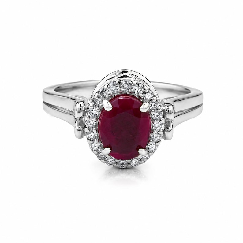May include: A silver-coloured ring featuring a central oval-cut deep red gemstone surrounded by a halo of small, clear, round-cut stones. The band is polished and smooth, with a split-shank design that connects to the central setting.