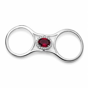 May include: A double-finger ring crafted from polished silver-toned metal, featuring a central oval-cut deep red gemstone surrounded by a halo of small, clear, sparkling stones. The design connects two circular bands with a central bridge.