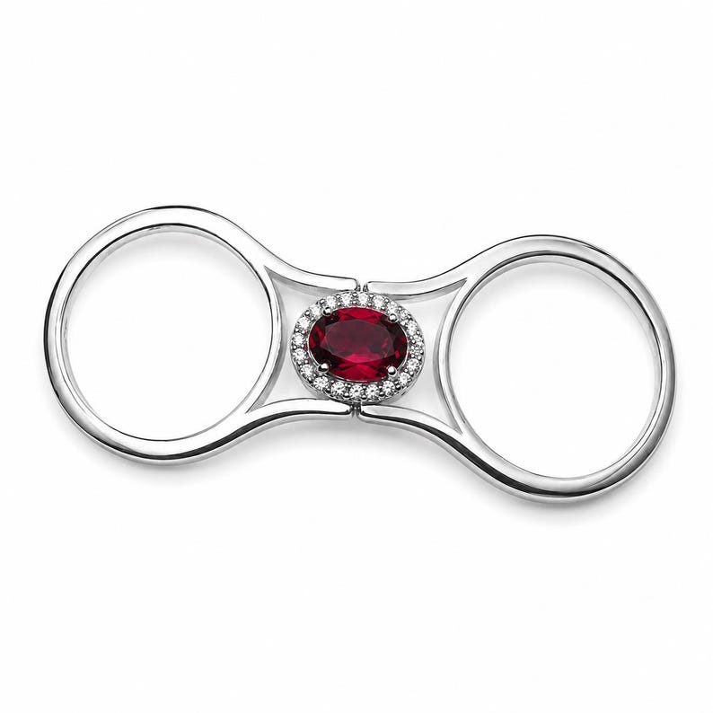 May include: A double-finger ring crafted from polished silver-toned metal, featuring a central oval-cut deep red gemstone surrounded by a halo of small, clear, sparkling stones. The design connects two circular bands with a central bridge.
