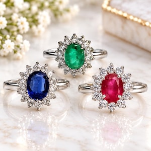 May include: Three rings with silver-toned bands, each featuring a central oval-cut gemstone surrounded by a halo of small round white stones. The gemstones are a deep blue sapphire, a vibrant green emerald, and a rich red ruby, all set in metal.