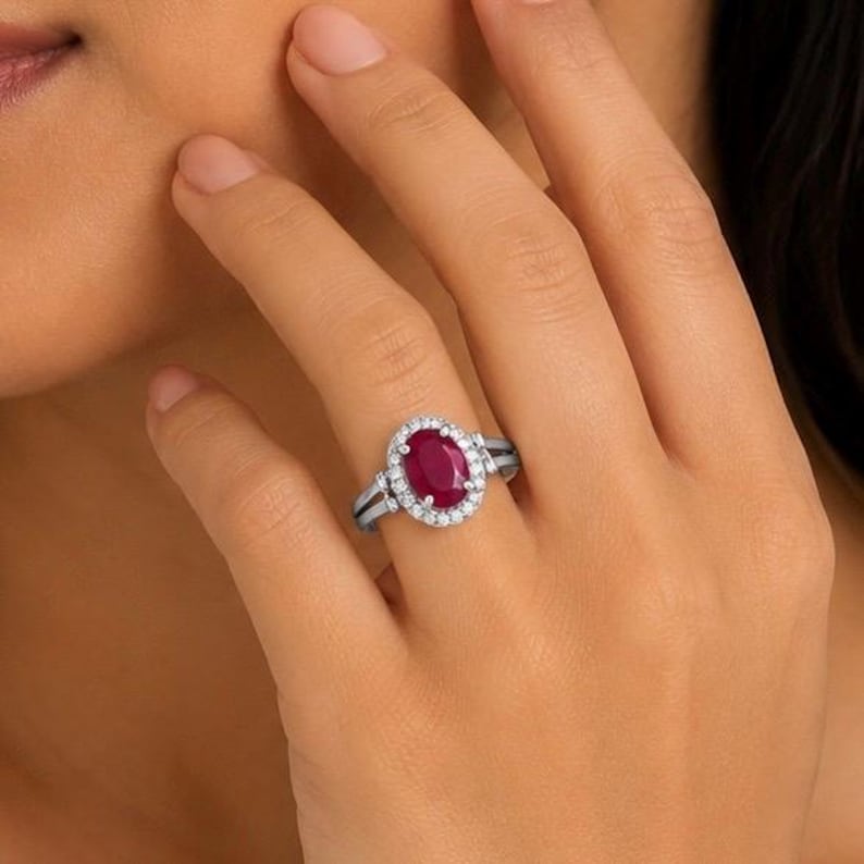 May include: A silver ring featuring a central oval-cut deep red ruby gemstone surrounded by a halo of small round white diamonds. The band is made of polished silver and has a split shank design that tapers towards the gemstone setting.
