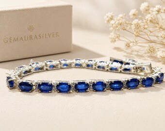 May include: A silver tennis bracelet featuring a series of oval-cut deep blue gemstones alternating with small clear stones. The bracelet is positioned in front of a beige box with the text GEMAURASILVER printed on the front in gold lettering.