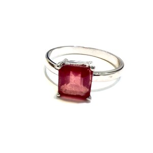 Puede incluir: Un anillo de plata con una gran gema rectangular de color rojo intenso. La gema es el punto focal, engastada en una banda sencilla y elegante. El anillo se muestra sobre un fondo blanco.