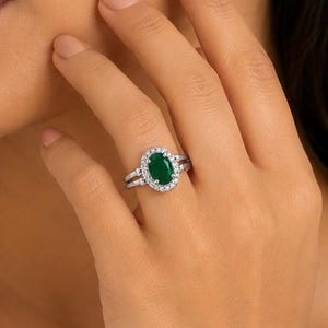 May include: A silver-toned ring featuring a central oval-cut green gemstone surrounded by a halo of small clear stones. The band is split into two sections on each side, leading up to the setting, creating a layered appearance on the finger.