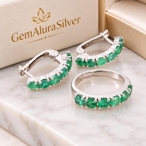 May include: A jewelry set consisting of a silver ring and two matching hoop earrings, each featuring a row of oval-cut green emerald gemstones. The items are displayed near a box labeled GemAlura Silver with a gold leaf logo design.