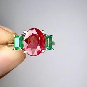 Op de afbeelding: Een zilverkleurige ring met een grote, ovaal geslepen rode robijn in het midden, geflankeerd door twee kleinere, rechthoekig geslepen groene smaragden. De stenen zijn gezet in een metalen zetting met pootjes voor een driestenen ontwerp.