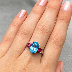 May include: A silver ring featuring a large, oval-cut, light blue gemstone flanked by two smaller, square-cut, magenta gemstones. The ring is set on a hand with manicured nails.