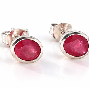 May include: A pair of stud earrings featuring oval-shaped, faceted red gemstones set in polished silver-toned metal bezels. Each earring includes a matching silver-toned metal post back, showcasing a clean, simple design with vibrant red stones.
