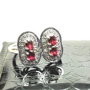 Peut inclure: Une paire de boucles d'oreilles ovales avec un sertissage argenté. Chaque boucle d'oreille présente deux pierres précieuses rouges vives entourées de petites pierres claires. Les boucles d'oreilles sont posées sur une surface sombre texturée.