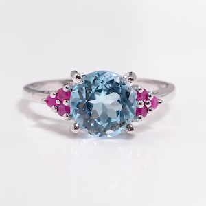 May include: A silver ring featuring a large, round, light blue gemstone as the centerpiece. Flanking the blue stone are clusters of smaller, vibrant pink gemstones. The ring is set against a white background.