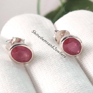Peut inclure: Une paire de boucles d'oreilles à clous avec des pierres précieuses ovales rouge foncé serties dans de l'argent. Les boucles d'oreilles sont présentées sur une surface blanche. Le texte "ShineGemsandJewllers" est visible.