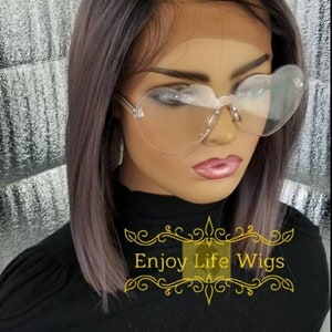 May include: A black bob wig with a lace front and a light purple ombre effect. The wig is styled straight and is worn on a mannequin head. The mannequin is wearing heart-shaped clear glasses and a black shirt with the text "Enjoy Life Wigs" in gold.