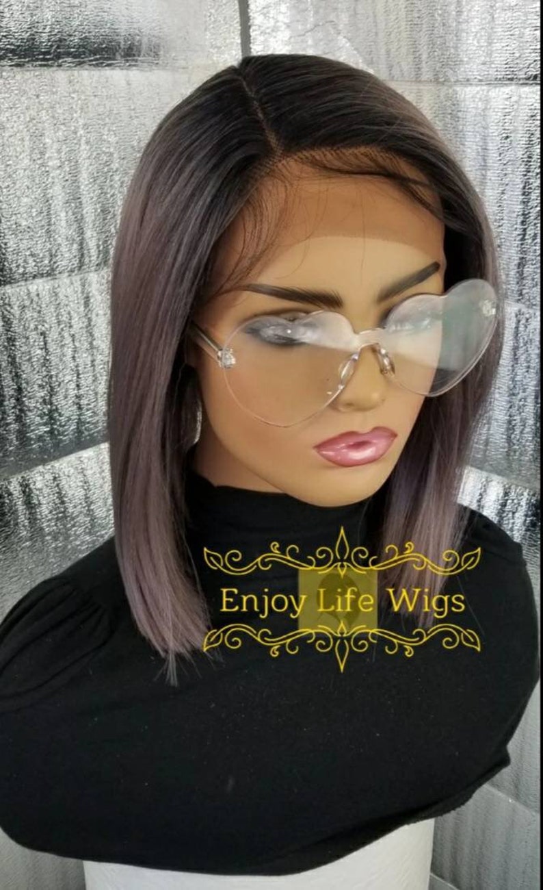 May include: A black bob wig with a lace front and a light purple ombre effect. The wig is styled straight and is worn on a mannequin head. The mannequin is wearing heart-shaped clear glasses and a black shirt with the text "Enjoy Life Wigs" in gold.
