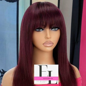 Può includere: Una parrucca lunga e liscia di colore bordeaux con frangia. La parrucca è acconciata su una testa di manichino. La parrucca è su uno sfondo rosa con un logo bianco che dice "ENJOY LIFE WIGS.COM".