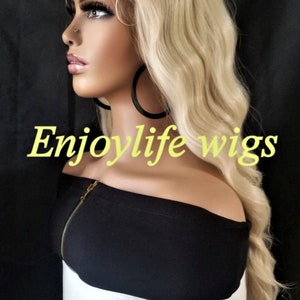 May include: A blonde wavy wig on a mannequin head. The wig is styled in a long, layered cut with loose waves. The mannequin is wearing a black off-the-shoulder top and large hoop earrings. The text "Enjoylife wigs" is visible in the image.