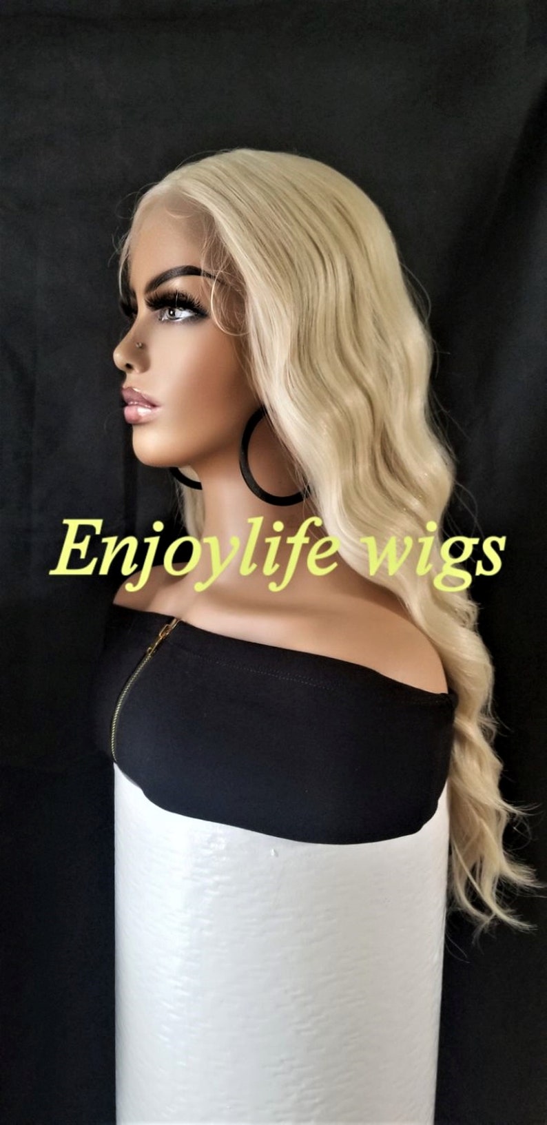 May include: A blonde wavy wig on a mannequin head. The wig is styled in a long, layered cut with loose waves. The mannequin is wearing a black off-the-shoulder top and large hoop earrings. The text "Enjoylife wigs" is visible in the image.