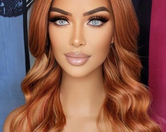 May include: A wig with long, wavy auburn hair with blonde highlights. The wig features a natural-looking hairline and is styled with soft waves. The person in the image has light blue eyes and full lips, with a full face of makeup.