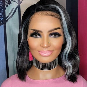 May include: A woman with a black and white wig, wearing a pink top and a silver choker. The wig has a side part with black hair and white highlights. She has long eyelashes and pink lipstick.