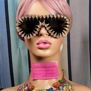 May include: A pair of black sunglasses with gold-toned metal frames and rhinestones. The sunglasses have a pointed design and are decorated with spikes. The sunglasses are being worn by a mannequin with pink hair and a colorful beaded necklace. The image also includes a pink card with the text "JUST4UDISCOUNT AUTHENTICATION CARD This product has been checked for quality."