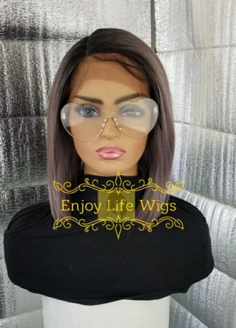 May include: A gray and black wig with a straight bob style. The wig is on a mannequin head wearing heart-shaped glasses. The text "Enjoy Life Wigs" is visible on the image.