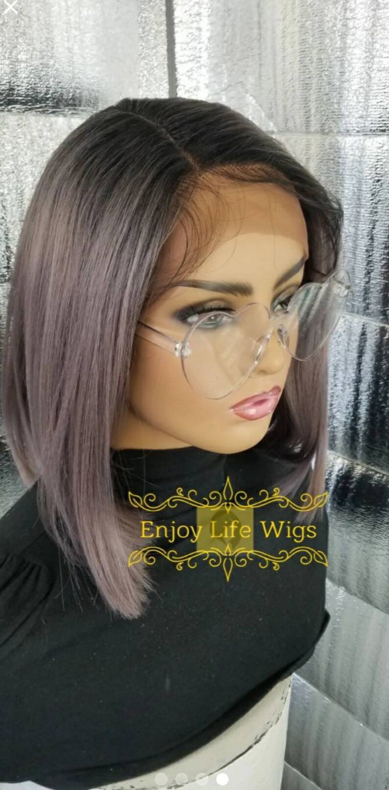 May include: A gray and purple ombre bob wig with a lace front. The wig is styled with a side part and is worn by a mannequin with heart-shaped glasses. The text "Enjoy Life Wigs" is visible on the mannequin's shirt.