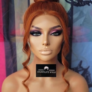 Puede incluir: Una peluca sintética de color cobre peinada en un moño alto con dos mechones largos, finos y rizados que enmarcan el rostro. La peluca presenta una línea de cabello frontal de encaje y un parche rectangular negro con el texto ENJOYLIFE WIGS.
