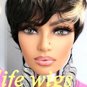 Puede incluir: Una peluca corta y entrecortada con cabello negro y rubio. La peluca está peinada con raya lateral y tiene un aspecto en capas. La peluca la lleva un maniquí con una camisa rosa y un collar de mariposa con joyas.