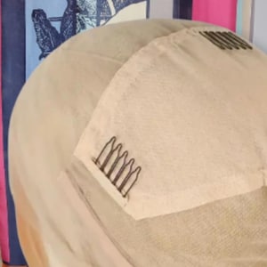 Pode incluir: Close-up de uma touca de peruca bege com pentes anexados para fixação segura. A touca é feita de um material fino e respirável, projetado para combinar com o tom de pele do usuário. Os pentes são marrons escuros.