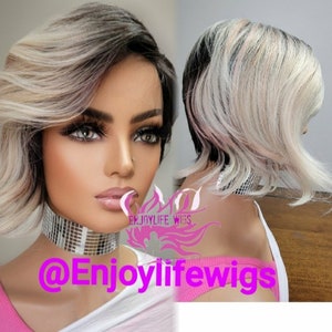 May include: A short, layered wig with a blend of black, blonde, and light pink tones. The wig features a side part and wavy texture. The image includes the text "Enjoylife Wigs" and a social media handle.
