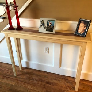May include: A light wood console table with a natural wood finish. The table has a rectangular top with a dark wood inlay. Two red candle holders with crystal accents are on the table. Two framed photos are on the table, one with a dragonfly and the other with a man. The table is in front of a white wall with a white baseboard.