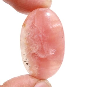 Puede incluir: Una piedra preciosa ovalada de color rosa y blanco con un patrón de remolino. La piedra tiene una superficie lisa y pulida y se sostiene entre dos dedos.