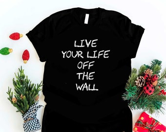 May include: Black t-shirt with white text that reads "Live your life off the wall". The shirt is surrounded by Christmas decorations, including a small pine tree, red and green ornaments, and a wrapped gift.