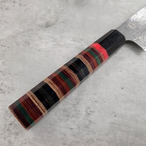 Puede incluir: Primer plano de un cuchillo de cocina con mango de madera negro y multicolor. La hoja es de acero inoxidable y tiene un patr&oacute;n de damasco.