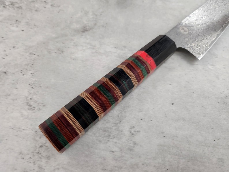 Puede incluir: Primer plano de un cuchillo de cocina con mango de madera negro y multicolor. La hoja es de acero inoxidable y tiene un patr&oacute;n de damasco.