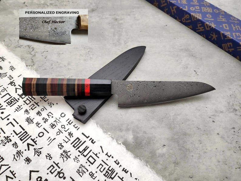 Puede incluir: Un cuchillo de cocina negro y gris con mango de madera y una grabaci&oacute;n personalizada que dice "Chef Hector". El cuchillo est&aacute; apoyado en una funda de cuero negro y un trozo de papel blanco con caracteres asi&aacute;ticos.