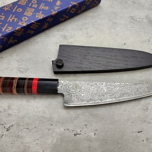 Puede incluir: Una funda de cuchillo de madera negra con un cuchillo de chef de estilo japon&eacute;s plateado con un mango con un patr&oacute;n rojo y negro. El cuchillo tiene una hoja de damasco.