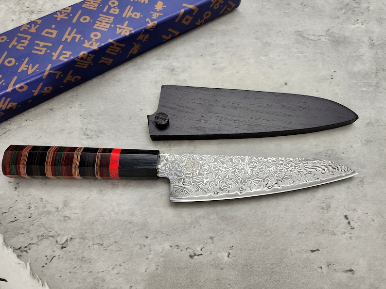 Puede incluir: Una funda de cuchillo de madera negra con un cuchillo de chef de estilo japon&eacute;s plateado con un mango con un patr&oacute;n rojo y negro. El cuchillo tiene una hoja de damasco.