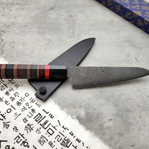 Puede incluir: Un cuchillo de cocina negro y gris con una hoja texturizada y un mango de madera con rayas rojas, negras y marrones. El cuchillo est&aacute; apoyado en una vaina de madera negra.