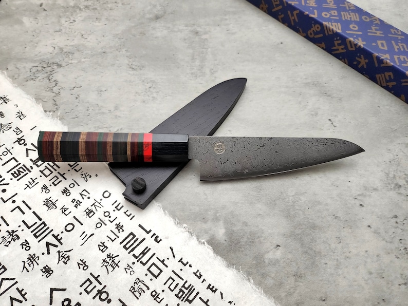 Puede incluir: Un cuchillo de cocina negro y gris con una hoja texturizada y un mango de madera con rayas rojas, negras y marrones. El cuchillo est&aacute; apoyado en una vaina de madera negra.