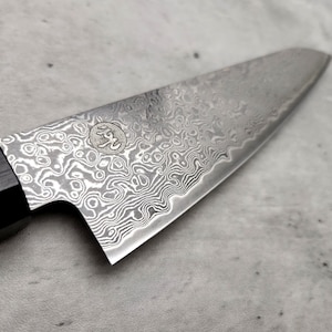 Puede incluir: Primer plano de un cuchillo de chef plateado con mango negro. La hoja tiene un patr&oacute;n ondulado distintivo y un peque&ntilde;o logotipo con caracteres japoneses.