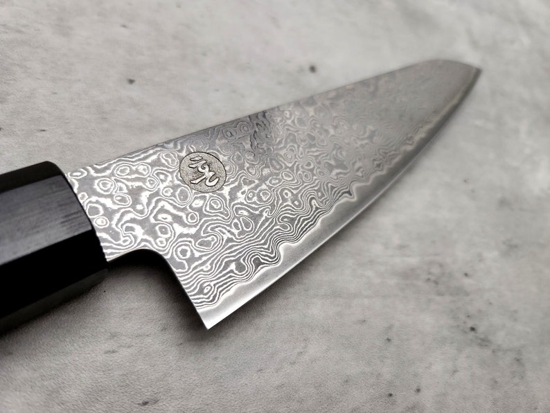Puede incluir: Primer plano de un cuchillo de chef plateado con mango negro. La hoja tiene un patr&oacute;n ondulado distintivo y un peque&ntilde;o logotipo con caracteres japoneses.