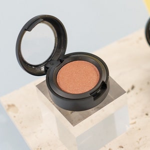 Könnte beinhalten: Ein runder, schwarzer Lidschatten-Behälter mit einem durchsichtigen Deckel. Der Lidschatten ist in einem schimmernden Pfirsich-Ton.
