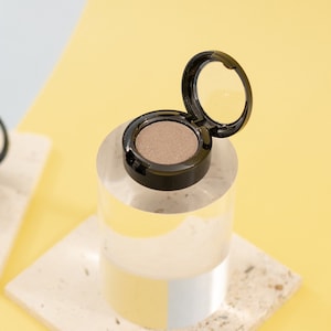 Könnte beinhalten: Zwei kleine runde Behälter mit Lidschatten, einer offen und einer geschlossen. Der offene Behälter befindet sich auf einem durchsichtigen Acrylzylinder, der auf einer hellbeigen, strukturierten Oberfläche steht. Der geschlossene Behälter befindet sich auf einer ähnlichen Oberfläche. Beide Behälter haben schwarze Deckel mit einem Spiegel im Inneren.