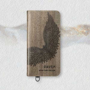 May include: A wooden phone case with a detailed raven wing design. The case is a light brown color with a dark brown wing illustration. The word "RAVEN" is printed below the wing, along with the words "WISDOM VISION PROTECTION". A metal loop is at the bottom.