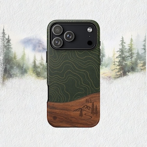 May include: A smartphone case featuring a dark green top section with gold topographic map lines and a dark brown wood grain bottom section. The wood portion displays an engraved mountain and pine tree graphic with the letters RLS.